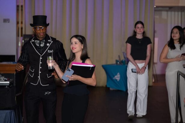 A man in a black suit and top hat stands beside a smiling woman holding a smartphone and a drink, while two other women stand in the background in a dimly lit indoor setting. South Florida Business & Wealth