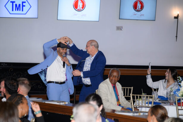 Two men in suits, one holding a microphone, dance together in front of a seated audience at an indoor event. People around them are smiling and watching, with one woman taking a photo. South Florida Business & Wealth
