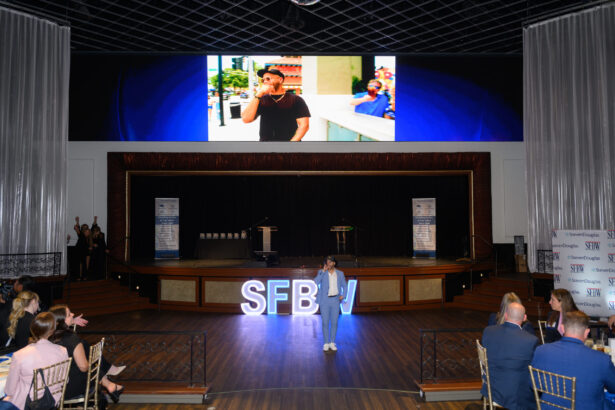 A person in a blue suit stands on stage in front of large lit-up "SFBW" letters, addressing an audience, with a big screen overhead displaying a man speaking on a phone outdoors. South Florida Business & Wealth
