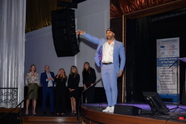 A man in a light blue suit and cap stands on stage holding a microphone, gesturing to the audience. Five people stand behind him, and a large banner with company information is visible on the right. South Florida Business & Wealth