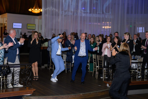 A group of people in formal attire enthusiastically dance and clap at an indoor event, with others seated at tables in the background. The room is decorated with draped curtains and chandeliers. South Florida Business & Wealth