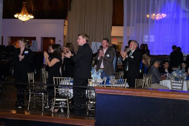 A group of people in formal attire stand and clap near tables with white tablecloths and decorative centerpieces in a dimly lit event venue with chandeliers and sheer curtains. South Florida Business & Wealth