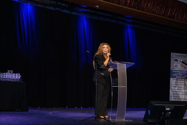 A woman in a black suit stands at a clear podium on a stage, speaking into a microphone. The stage is dimly lit with blue lights, and a banner and table with trophies are visible in the background. South Florida Business & Wealth