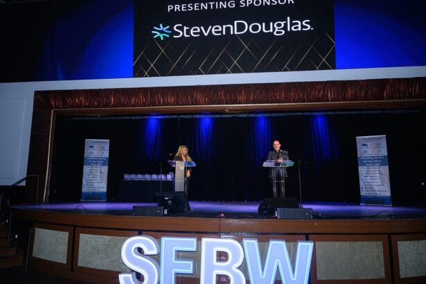 Two people stand at podiums on a stage with blue lighting. Behind them, a large screen displays “Presenting Sponsor StevenDouglas.” Large illuminated letters spell “SEBW” at the front of the stage. South Florida Business & Wealth