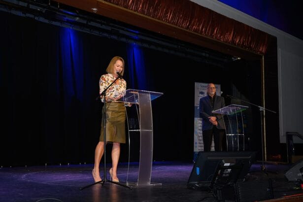 A woman stands at a clear podium speaking into a microphone on stage, while a man stands at another podium in the background. Both are dressed formally, with stage lights illuminating them against a dark backdrop. South Florida Business & Wealth