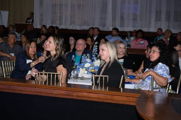 A group of people sit at round tables, smiling and attentively watching an event. The room is decorated with blue and white floral centerpieces, and most attendees are dressed in business or semi-formal attire. South Florida Business & Wealth