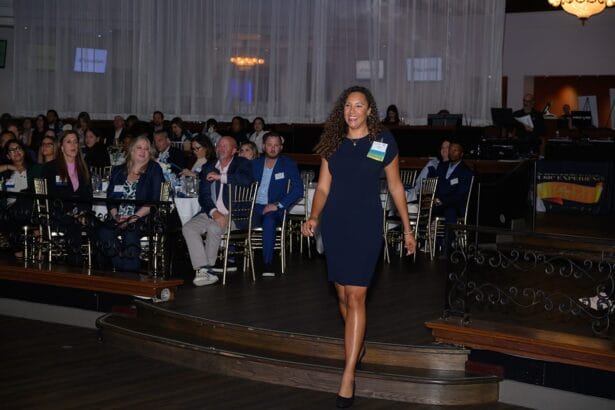 A woman in a navy dress with a name tag walks on stage at a formal event, smiling, while a seated audience watches her in a decorated venue with tables and curtains. South Florida Business & Wealth