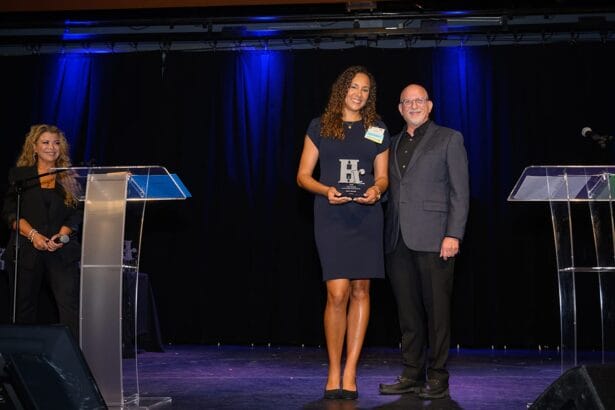 A woman holding an award stands beside a smiling man on stage, both facing the camera. Another woman stands at a podium on the left. The background is dark with blue lighting. South Florida Business & Wealth