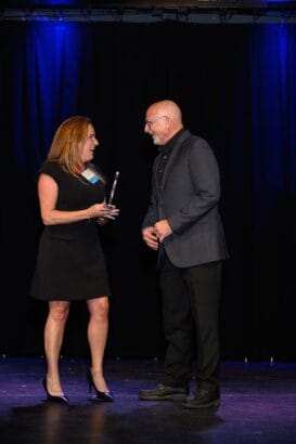 A woman in a black dress hands an award to a man in a dark suit on a stage with blue lighting and a black curtain backdrop. Both are smiling and engaged in conversation. South Florida Business & Wealth