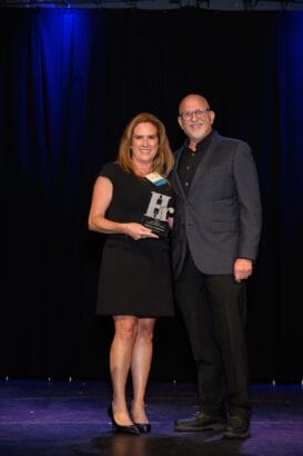 A woman in a black dress holding an award stands next to a man in a suit, both smiling, on a stage with a black curtain and blue lighting in the background. South Florida Business & Wealth