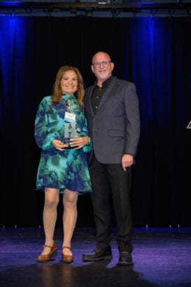 A woman in a blue-green dress holds a trophy and stands next to a man in a dark suit jacket. Both are smiling on a stage with dark curtains and blue lighting in the background. South Florida Business & Wealth