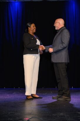 Two people dressed in business attire stand on a stage with a black curtain background, shaking hands and smiling at each other under blue stage lights. South Florida Business & Wealth