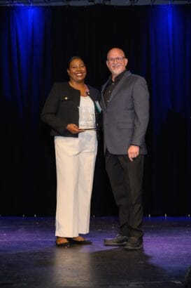 Two people stand smiling on a stage with a dark backdrop and blue lighting. The person on the left holds a clear award trophy, and both are dressed in business attire. South Florida Business & Wealth