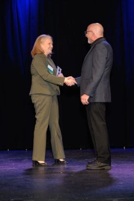 A woman and a man in business attire stand on a stage, shaking hands. The woman holds a clear award and both are smiling. A dark curtain and blue stage lighting provide the background. South Florida Business & Wealth