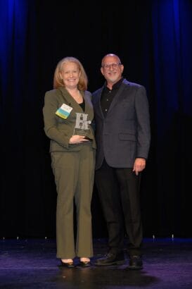 A woman in a suit holding a trophy and smiling stands next to a man in a dark suit, both facing the camera on a stage with a dark curtain backdrop. South Florida Business & Wealth