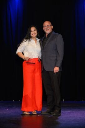A woman in a white blouse and bright red pants stands next to a man in a dark suit on a stage, both smiling, with the woman holding an award. The background is black curtains with blue lighting above. South Florida Business & Wealth