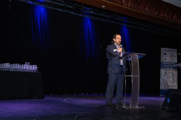 A man in a suit speaks at a clear podium on a stage with blue lighting. Stacks of small boxes sit on a table to the side, and a standing banner is visible in the background. South Florida Business & Wealth