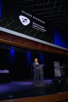 A man stands at a clear podium speaking on stage beneath a large screen that reads "Silver Sponsor: Recovery Unplugged Behavioral Health" with a logo. The stage is lit with blue lighting. South Florida Business & Wealth