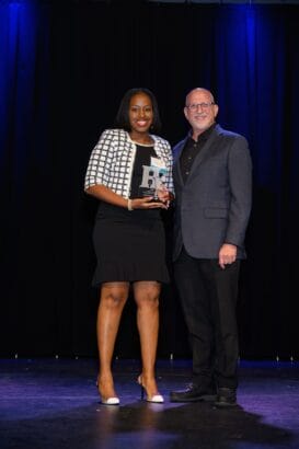 A woman in a black dress and checkered jacket holds a glass award while standing next to a smiling man in a gray suit. They are on a stage with a dark curtain backdrop and blue lights above. South Florida Business & Wealth
