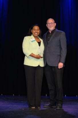 A woman in a light green blazer holds an award while standing next to a man in a gray suit. They are smiling and posing on a stage with a dark curtain and blue lighting in the background. South Florida Business & Wealth