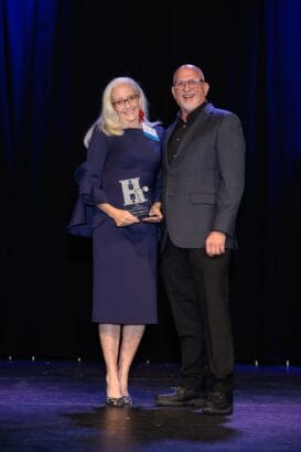 An older woman in a dark blue dress holding an award stands beside a smiling man in a blazer and dark pants. They pose together on a stage with a dark curtain background. South Florida Business & Wealth