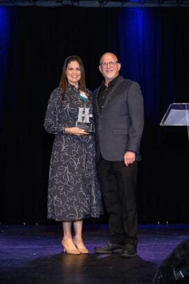 A woman in a patterned dress holds an award and stands next to a man in a dark suit on a stage with dark curtains and blue lights. Both are smiling and facing the camera. South Florida Business & Wealth