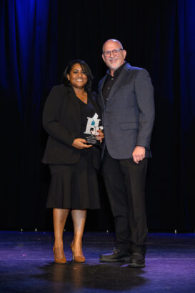 A woman holding an award and a man stand smiling together on a stage with a black curtain backdrop. Both are dressed in formal attire. South Florida Business & Wealth