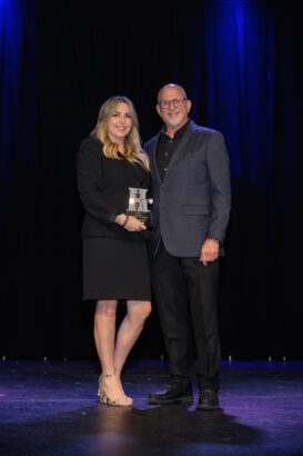 A woman in a black dress holding an award stands next to a man in a dark suit on a stage with a black curtain and blue lights in the background. South Florida Business & Wealth