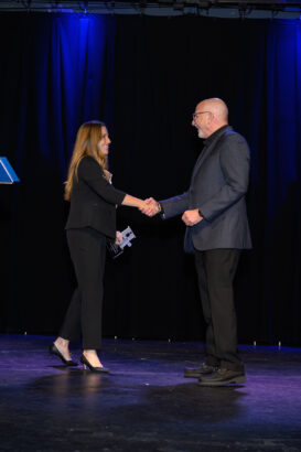 A woman and a man, both dressed in business attire, shake hands on a stage with a dark curtain and blue lighting in the background. The woman is holding some papers. South Florida Business & Wealth
