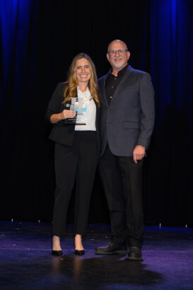 Two people stand on a stage in business attire, smiling at the camera. The woman on the left holds a clear award. The background is dark with blue light accents. South Florida Business & Wealth