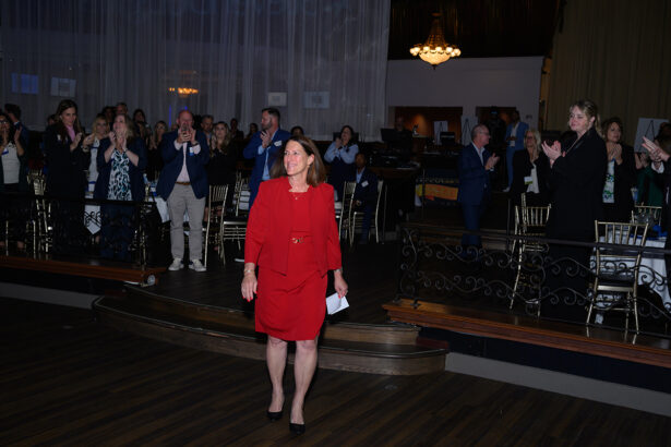 A woman in a red suit stands on a stage with people in the audience standing and applauding her. The event appears formal with elegant decor, dim lighting, and round tables. South Florida Business & Wealth