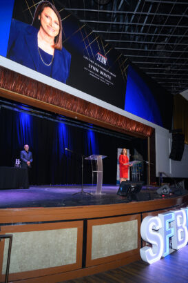 A woman stands at a podium on stage during an award ceremony, with a large screen above displaying her image and the text “SFBW Lifetime Achievement Award: Lynn White.” An audience member stands nearby. South Florida Business & Wealth