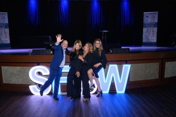 Four people pose cheerfully in front of large illuminated letters spelling "SHOW" on a stage. The background features blue lighting and event banners. The group appears happy and dressed in formal or semi-formal attire. South Florida Business & Wealth