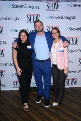 Three people pose and smile in front of a step-and-repeat backdrop with "SFBW" and "StevenDouglas" logos. The man in the center wears a blue suit, while the two women on either side wear business attire. South Florida Business & Wealth