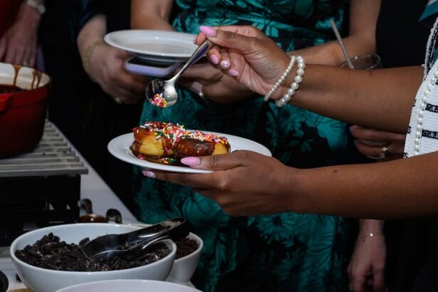 A person holds a plate with a chocolate-covered pastry topped with rainbow sprinkles, using a spoon to add more sprinkles. Several bowls with toppings and other people are visible in the background. South Florida Business & Wealth