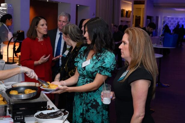 A group of people in business attire stand in line at a buffet, with one person serving food onto plates. The atmosphere appears to be a formal social or networking event. South Florida Business & Wealth