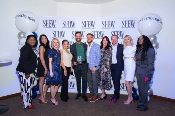 A group of ten people stands smiling in front of an SFBW step-and-repeat backdrop with StevenDouglas balloons on either side, at what appears to be an event or awards ceremony. South Florida Business & Wealth