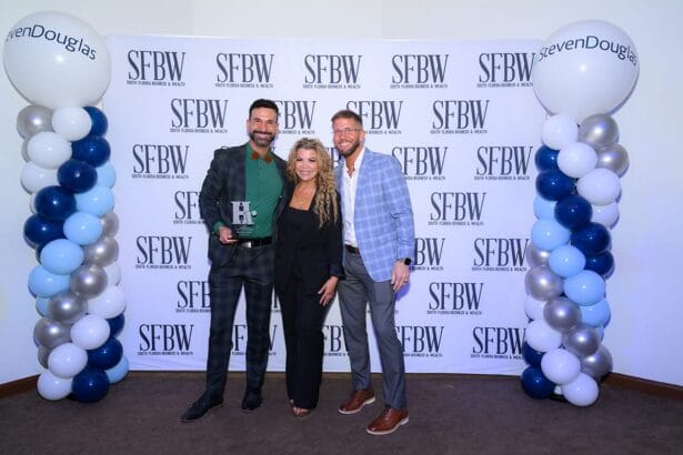 Three people stand smiling in front of an SFBW step-and-repeat banner flanked by two balloon columns. The person on the left holds an award. All are dressed in business attire. South Florida Business & Wealth