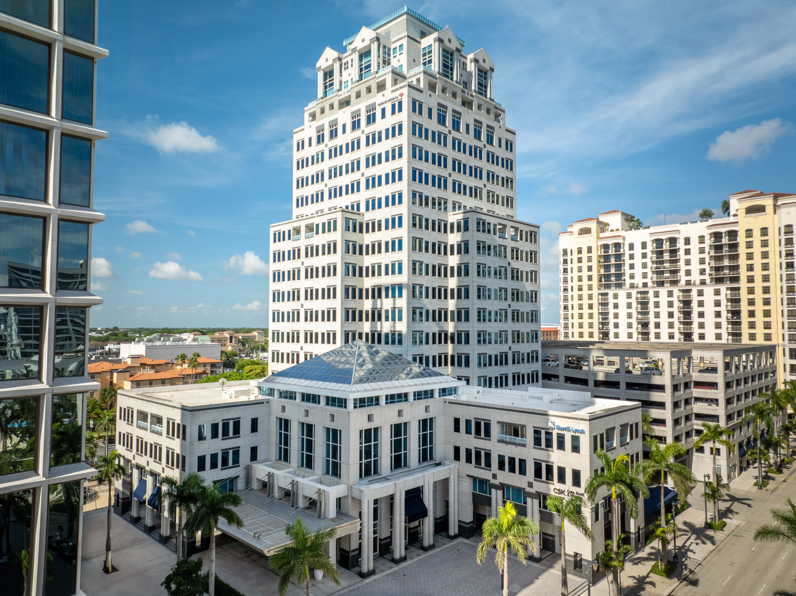 A tall, white, multi-story office building with a unique rooftop, surrounded by palm trees and smaller buildings, set against a blue sky with scattered clouds. South Florida Business & Wealth