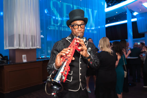 A musician in a black and white suit and top hat plays a red saxophone at an indoor event, with people mingling and blue lighting in the background. South Florida Business & Wealth