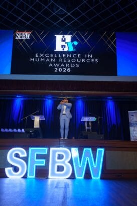 A person in a blue suit speaks on stage at the "Excellence in Human Resources Awards 2026" event, with a large "SFBW" light sign in front and a digital screen above displaying the event name. South Florida Business & Wealth