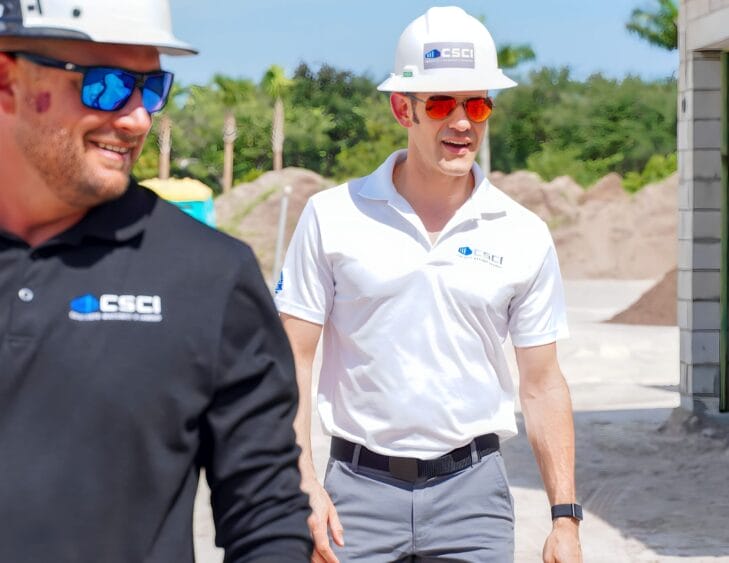 Two men wearing hard hats and CSCI-branded shirts smile and walk at a sunny construction site, with piles of dirt, trees, and a building visible in the background. South Florida Business & Wealth