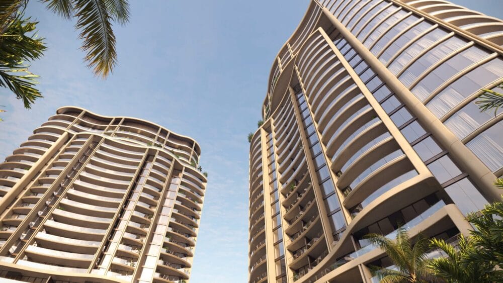 Two modern high-rise buildings with curved balconies, viewed from below against a blue sky. Palm trees frame the scene, adding a tropical atmosphere. South Florida Business & Wealth