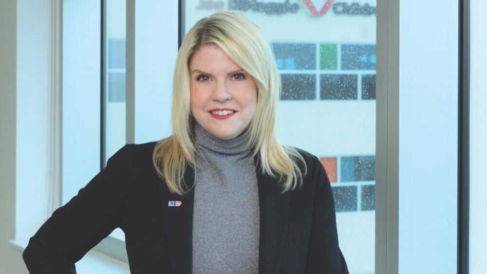 A woman with blonde hair, wearing a gray turtleneck and black blazer, smiles while standing indoors by large windows with a blurred building in the background. South Florida Business & Wealth