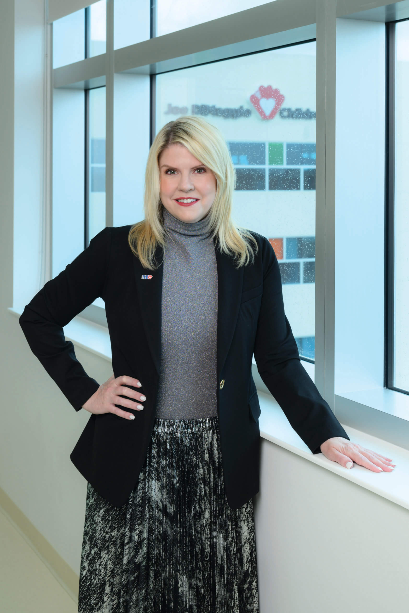 A woman with blonde hair, wearing a black blazer, gray turtleneck, and patterned skirt, stands by a window with one hand on her hip and the other resting on the ledge. A building with a heart logo is visible outside. South Florida Business & Wealth