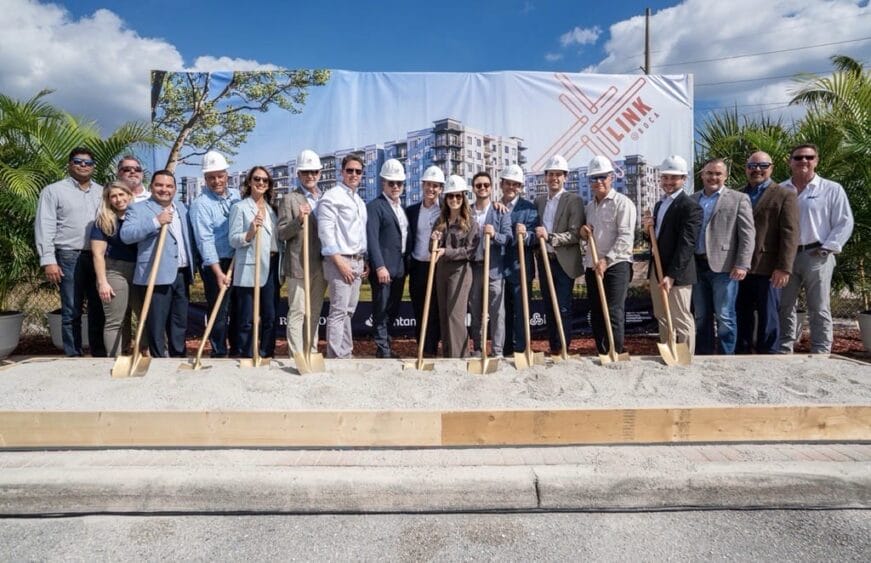 A group of people in business attire and hard hats stand in front of a construction site banner, holding shovels and posing for a groundbreaking ceremony on a sunny day. South Florida Business & Wealth