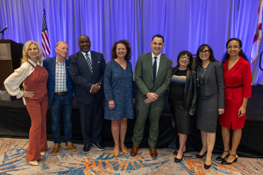 A group of eight people, dressed in business and semi-formal attire, stand smiling together for a photo in front of a stage with blue curtains, an American flag, and a Florida flag. South Florida Business & Wealth