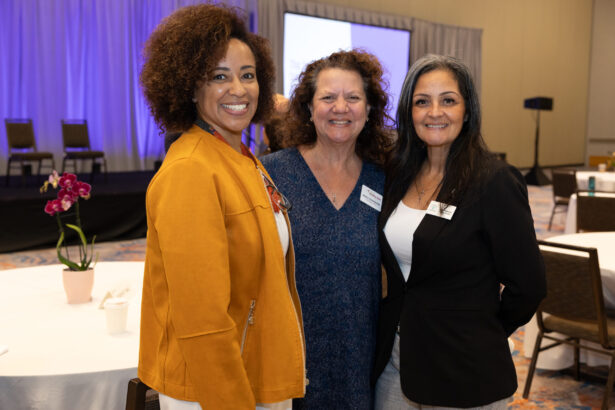 Three women stand together smiling at a conference or event, with round tables, chairs, and a stage with a purple backdrop visible in the background. South Florida Business & Wealth