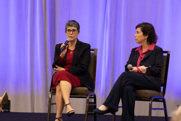 Two women sit on chairs on a stage with a purple curtain behind them. The woman on the left holds a microphone and speaks, while the woman on the right looks at her, also holding a microphone. Both wear business attire. South Florida Business & Wealth