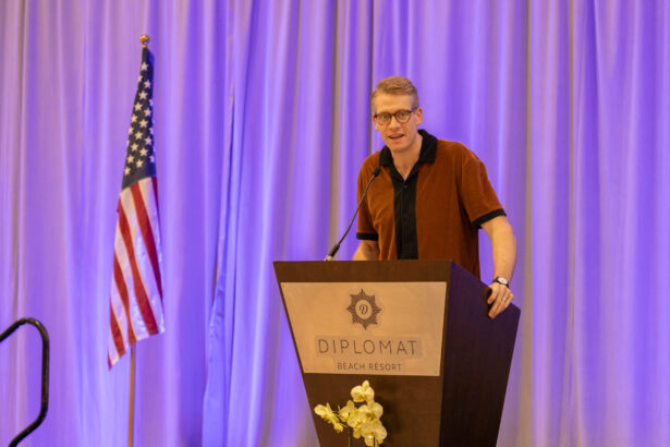 A man in glasses speaks at a podium labeled "Diplomat Beach Resort" with an American flag and purple curtains in the background. South Florida Business & Wealth